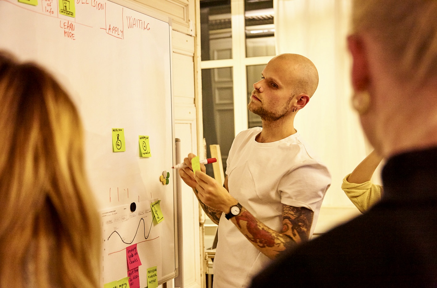 Daniel Majoros at a whiteboard during a workshop, adding a sticky note to a user-journey map covered in hand-drawn sketches and notes.